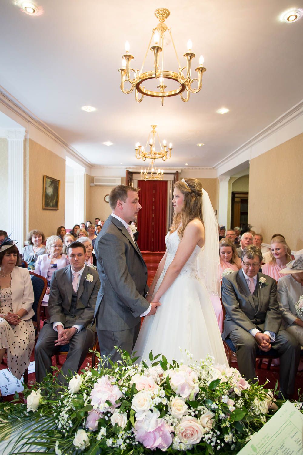 Elegant wedding ceremony in a formal room with crystal chandeliers and beautiful floral arrangements in soft pink and white.