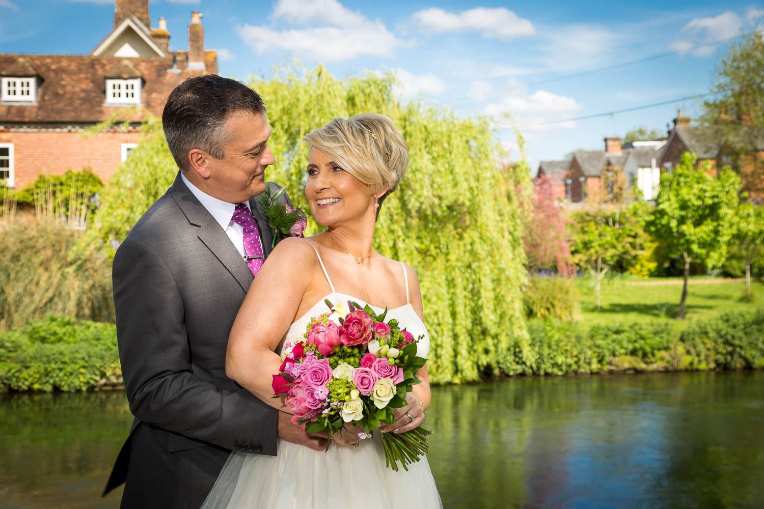 A newlywed couple stands by a tranquil pond with weeping willows and pink floral bouquet on their wedding day.