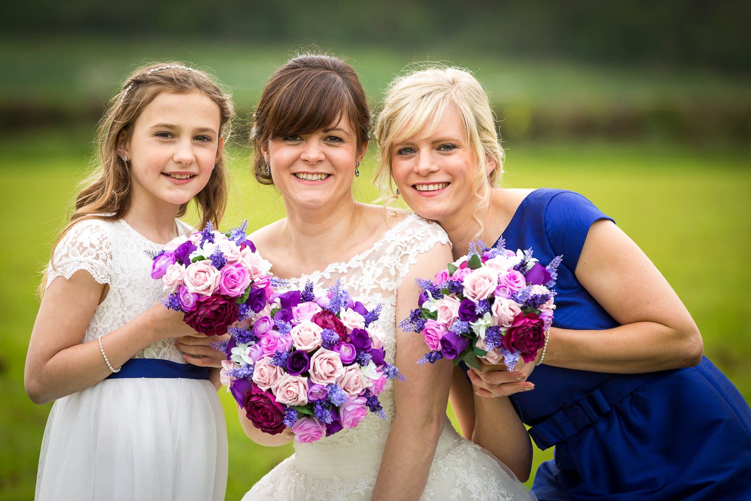 Three bridal party members pose together outdoors holding vibrant purple and pink flower bouquets.