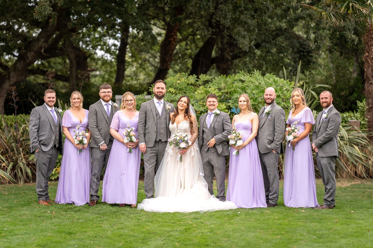 Wedding party posing together with bridesmaids in lavender gowns and groomsmen in grey suits outdoors.