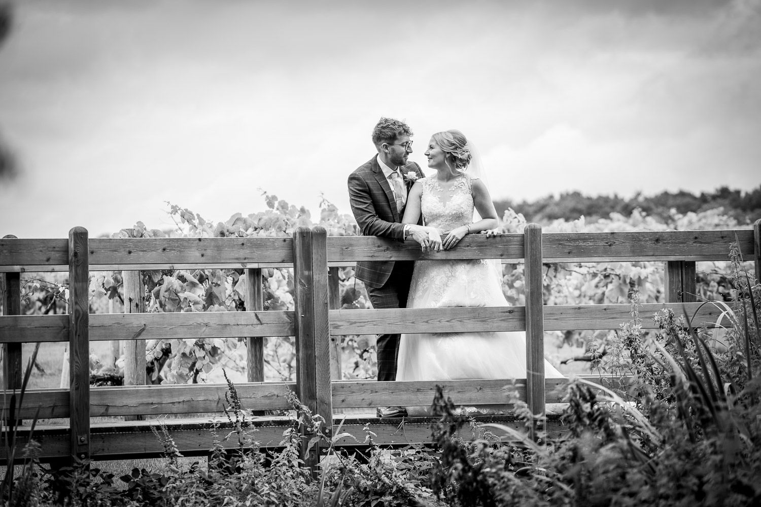 A romantic black and white photograph of a couple standing against a wooden fence in a scenic countryside setting.