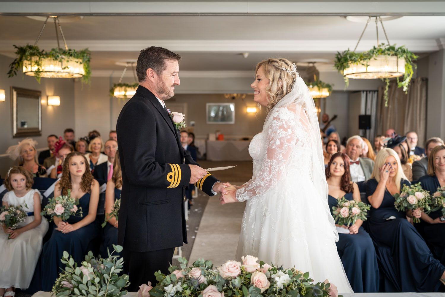A couple exchanges vows at a formal wedding ceremony with uniformed groom and bride in lace dress, surrounded by guests.