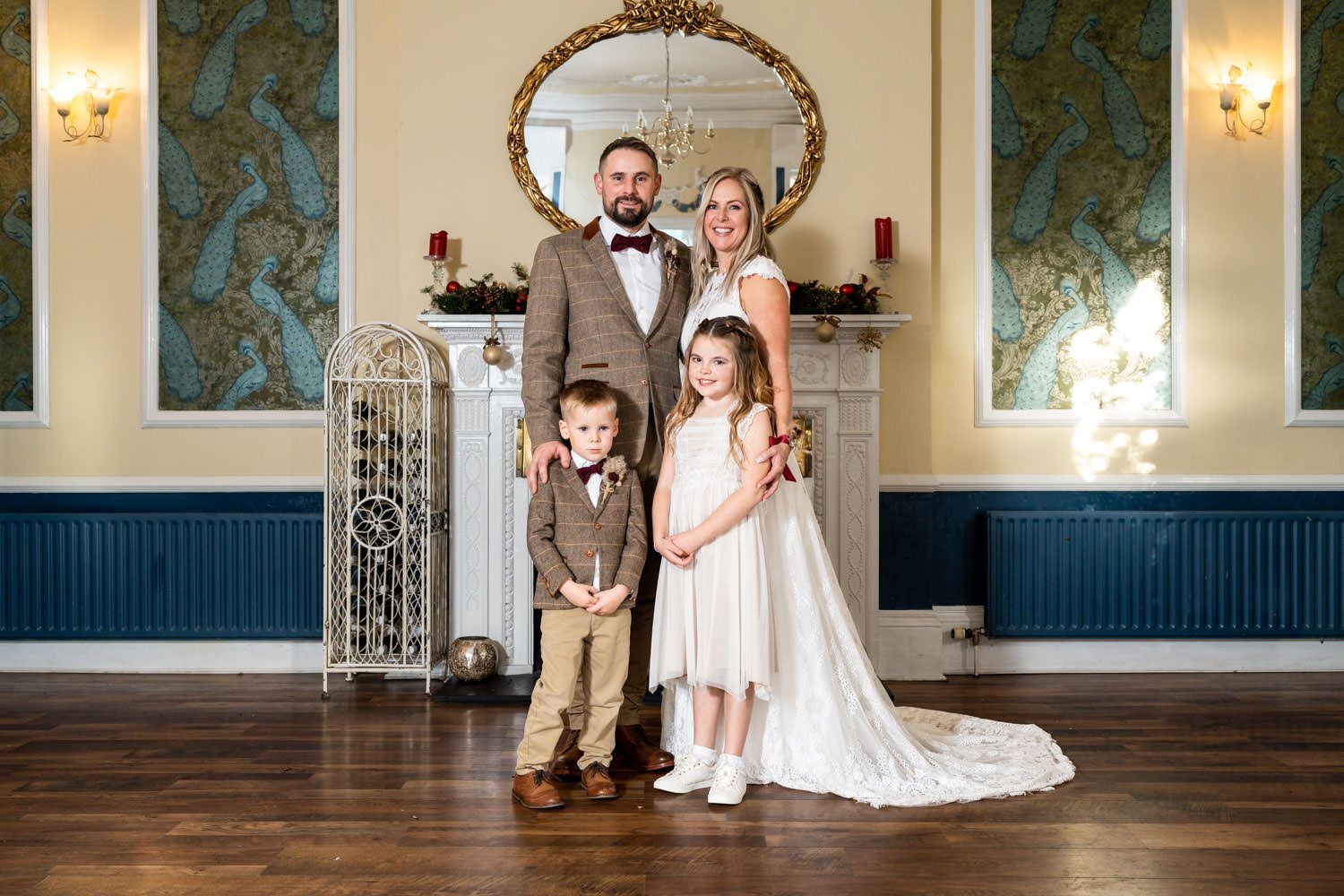A formal group stands in an elegant room with teal wallpaper and ornate mirror, dressed in wedding attire.
