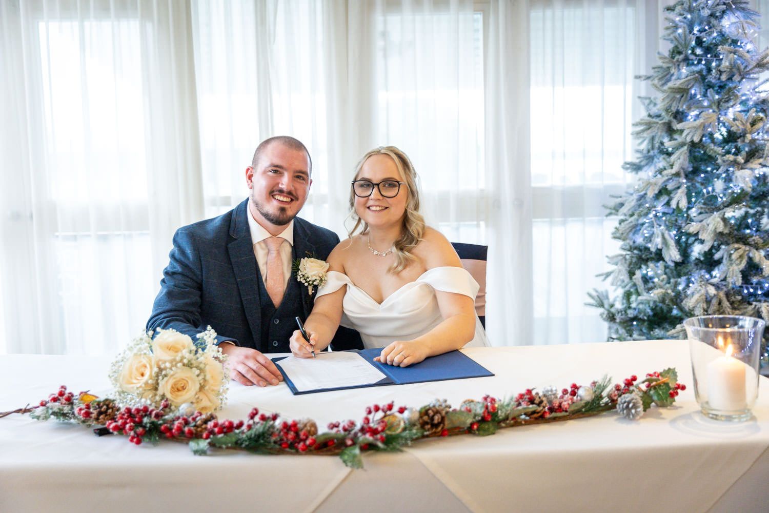 Couple sits at decorated table signing wedding documents during winter ceremony with Christmas tree in background.