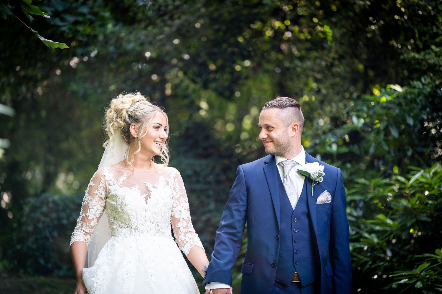 A newlywed couple in wedding attire holding hands outdoors with greenery in the background.