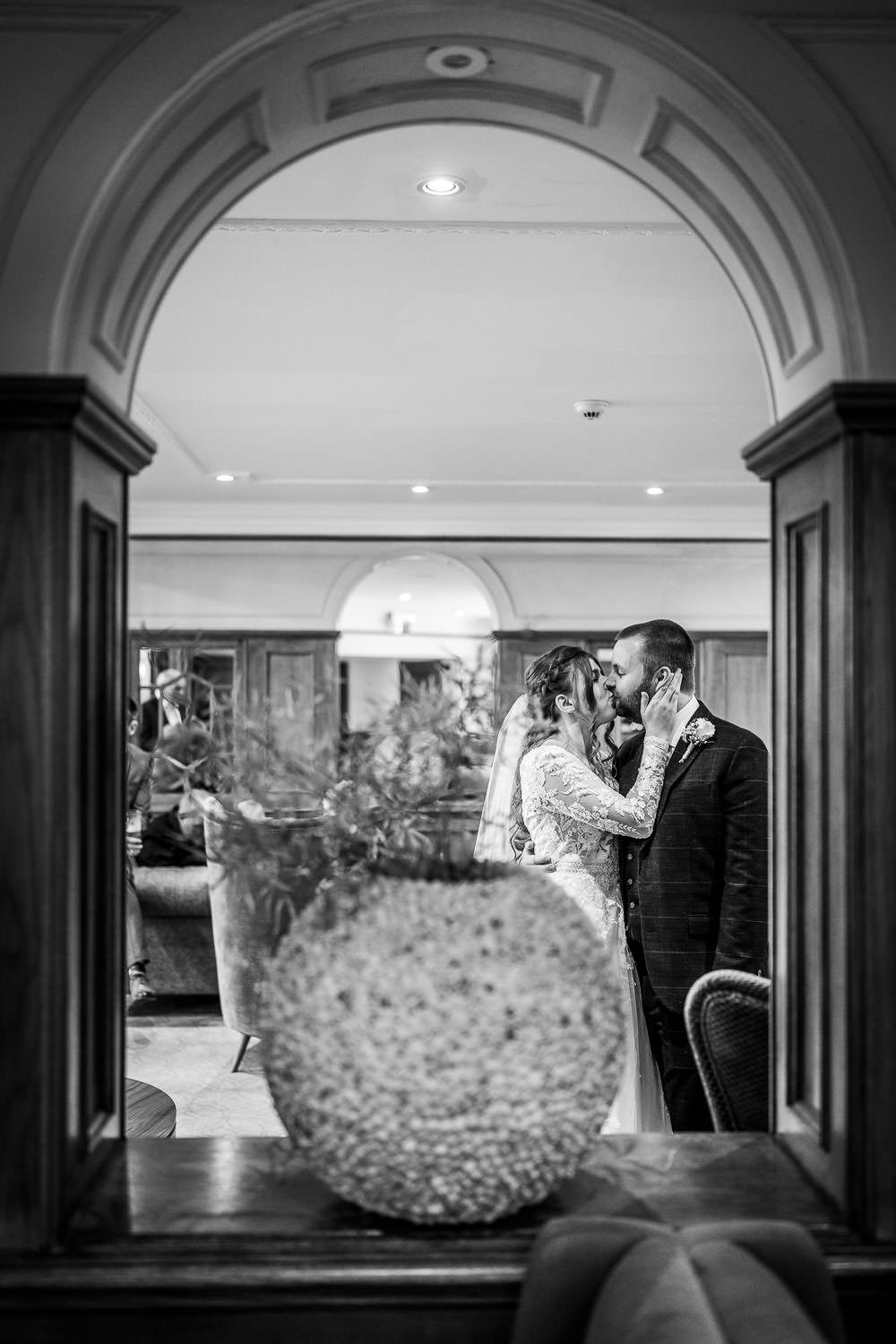 Black and white photograph showing a romantic moment viewed through an elegant arched doorway in an upscale venue.