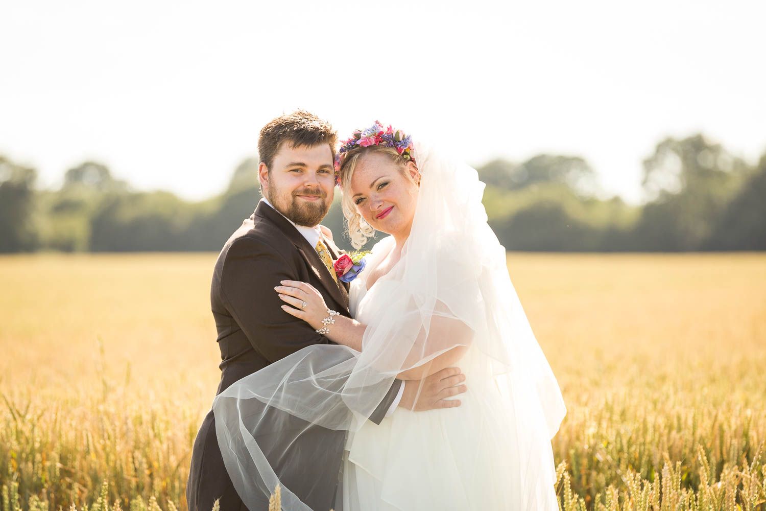 A happy wedding couple embraces in a golden wheat field at sunset while the bride's veil flows in the breeze.