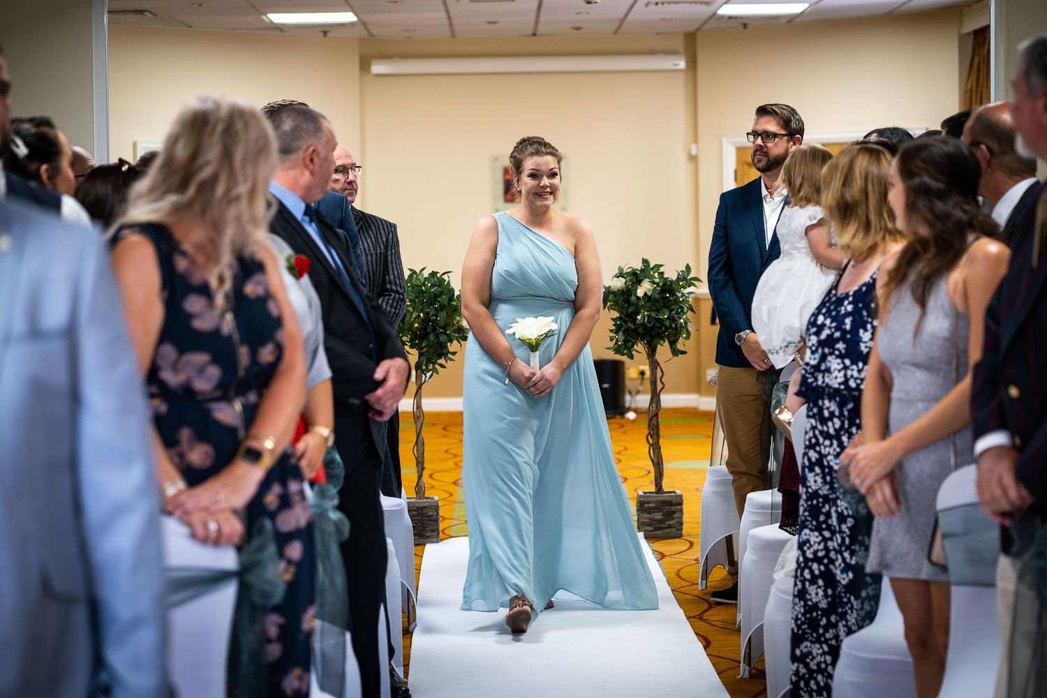Wedding ceremony with attendees watching as bridesmaid in light blue gown walks down aisle in indoor venue.