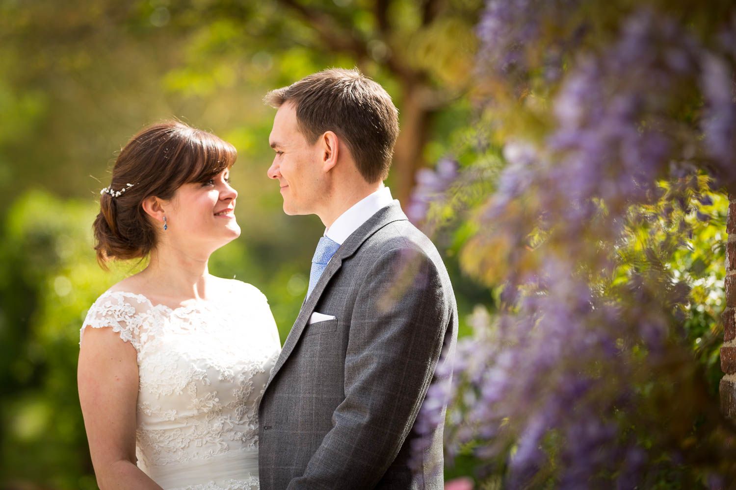 A romantic wedding couple share a tender moment in a garden with purple flowers in the background.