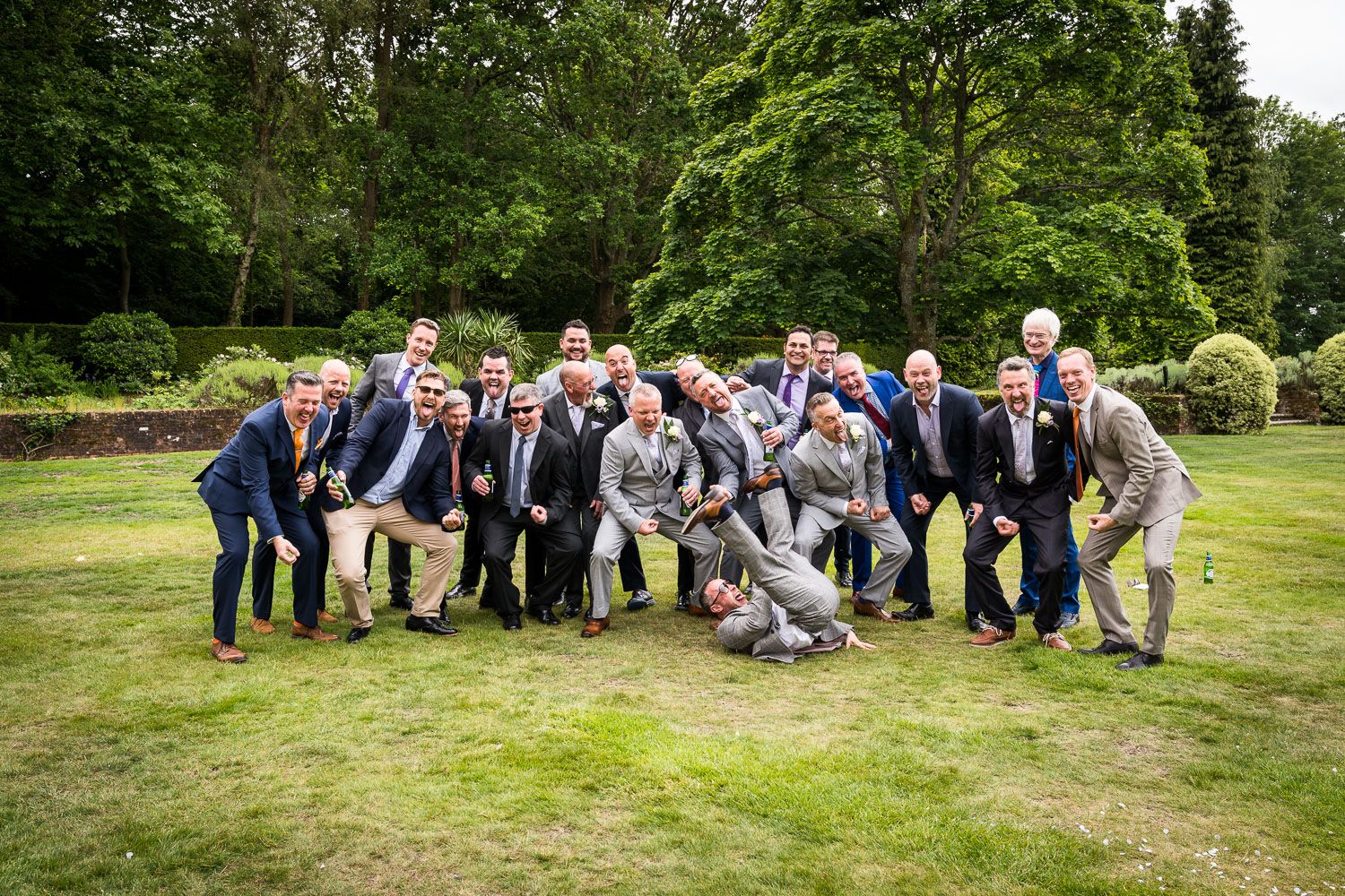 A group in formal attire poses together playfully on a grassy lawn surrounded by green trees at an outdoor event.