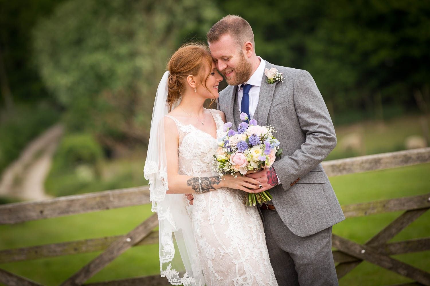A bride and groom share a tender moment by a rustic wooden fence, with the bride holding a purple and white bouquet.