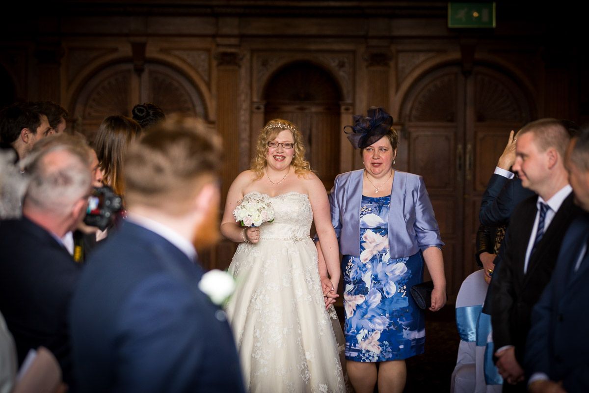 Bride in white lace wedding gown enters church ceremony space accompanied by woman in blue floral dress.