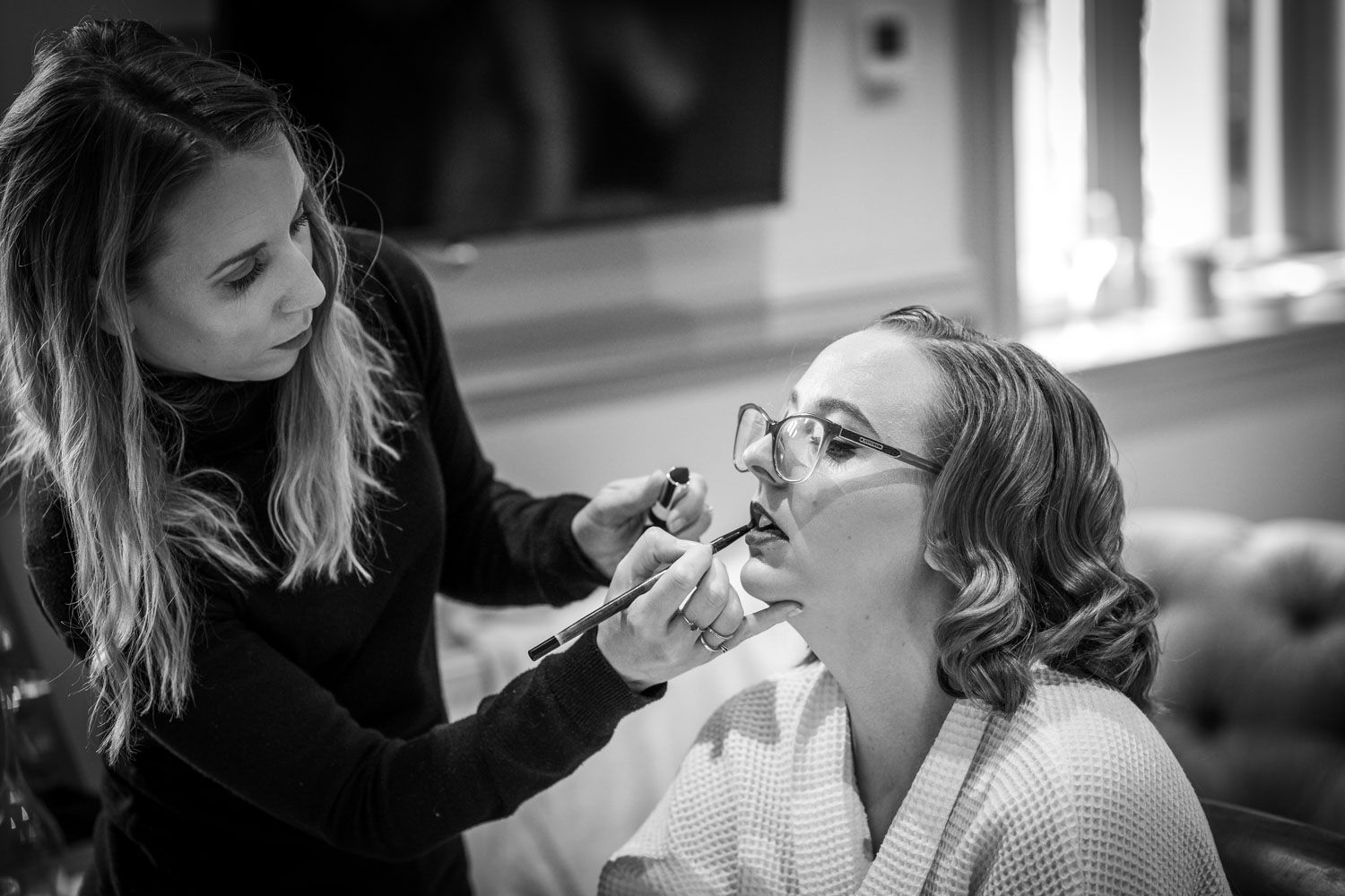 Black and white image of a makeup artist applying lipstick to someone wearing glasses and a robe during beauty prep.