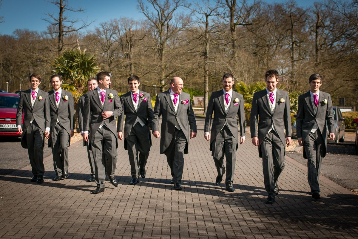 A group of groomsmen in matching gray suits with pink accessories walk together on a paved path.