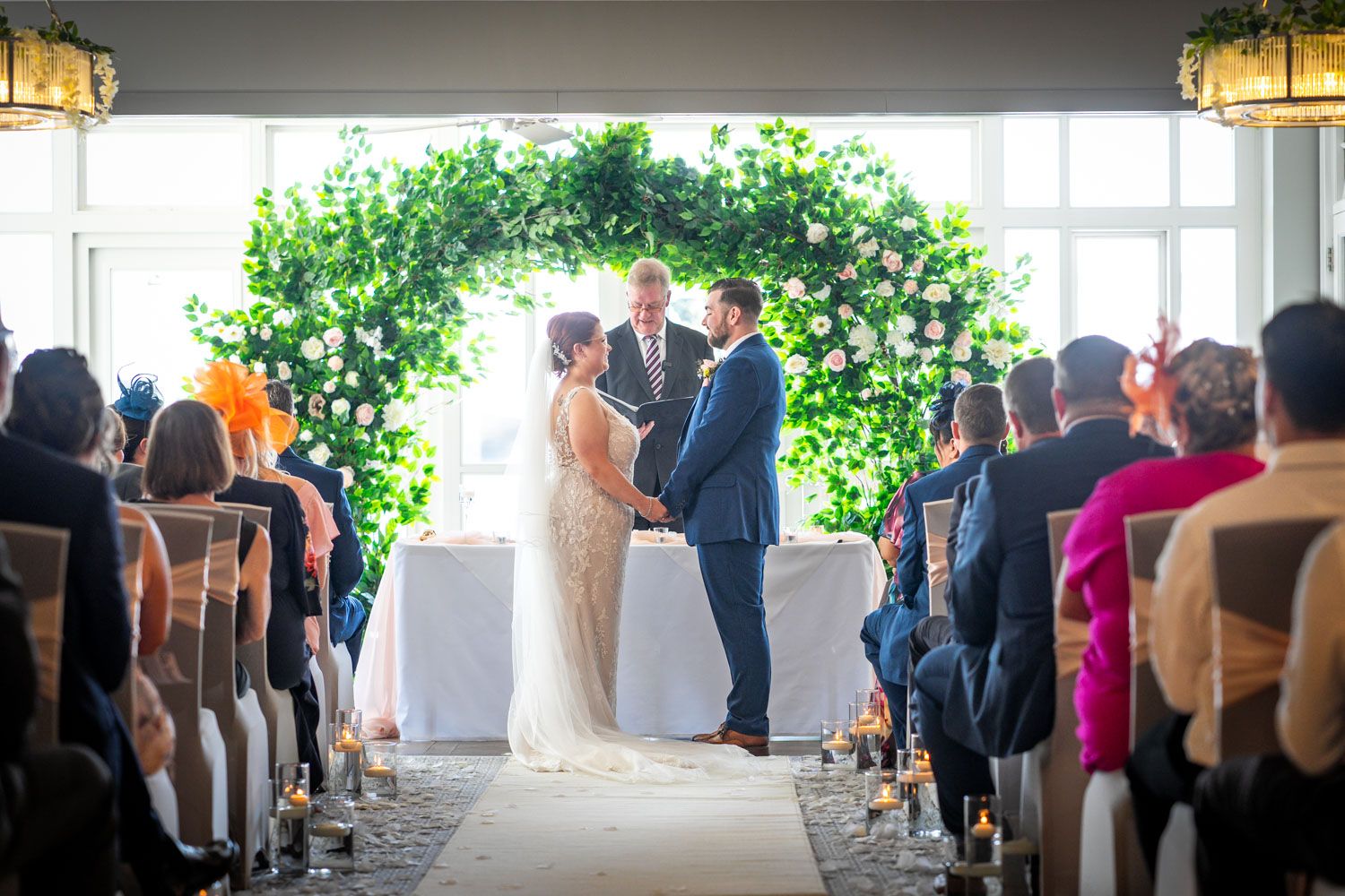 Wedding ceremony taking place under a lush green floral arch with guests seated in rows during an indoor celebration.