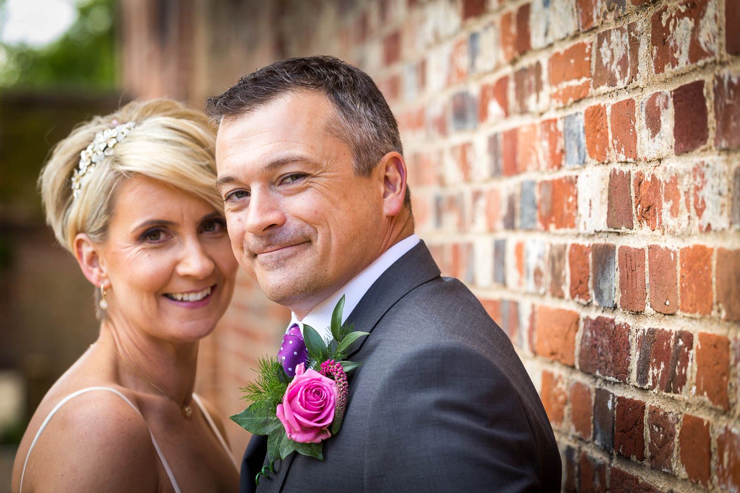 A smiling couple poses against a rustic brick wall during a wedding ceremony, with a pink rose boutonniere accent.