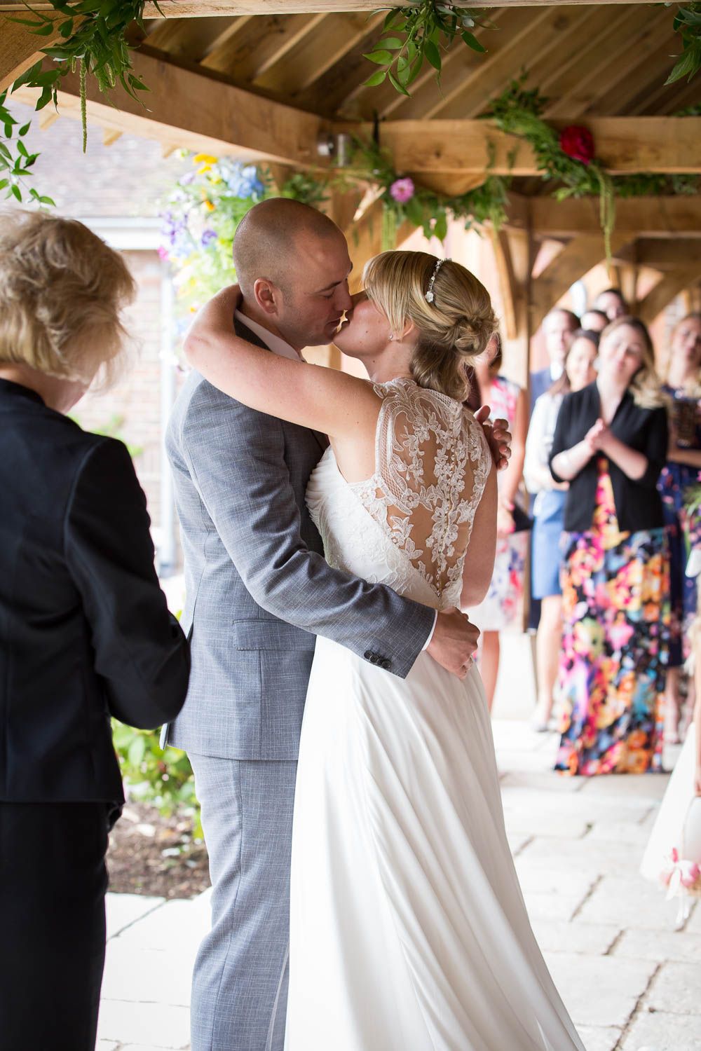 A couple shares their first kiss at an outdoor wedding ceremony while guests look on from under a rustic wooden pavilion.
