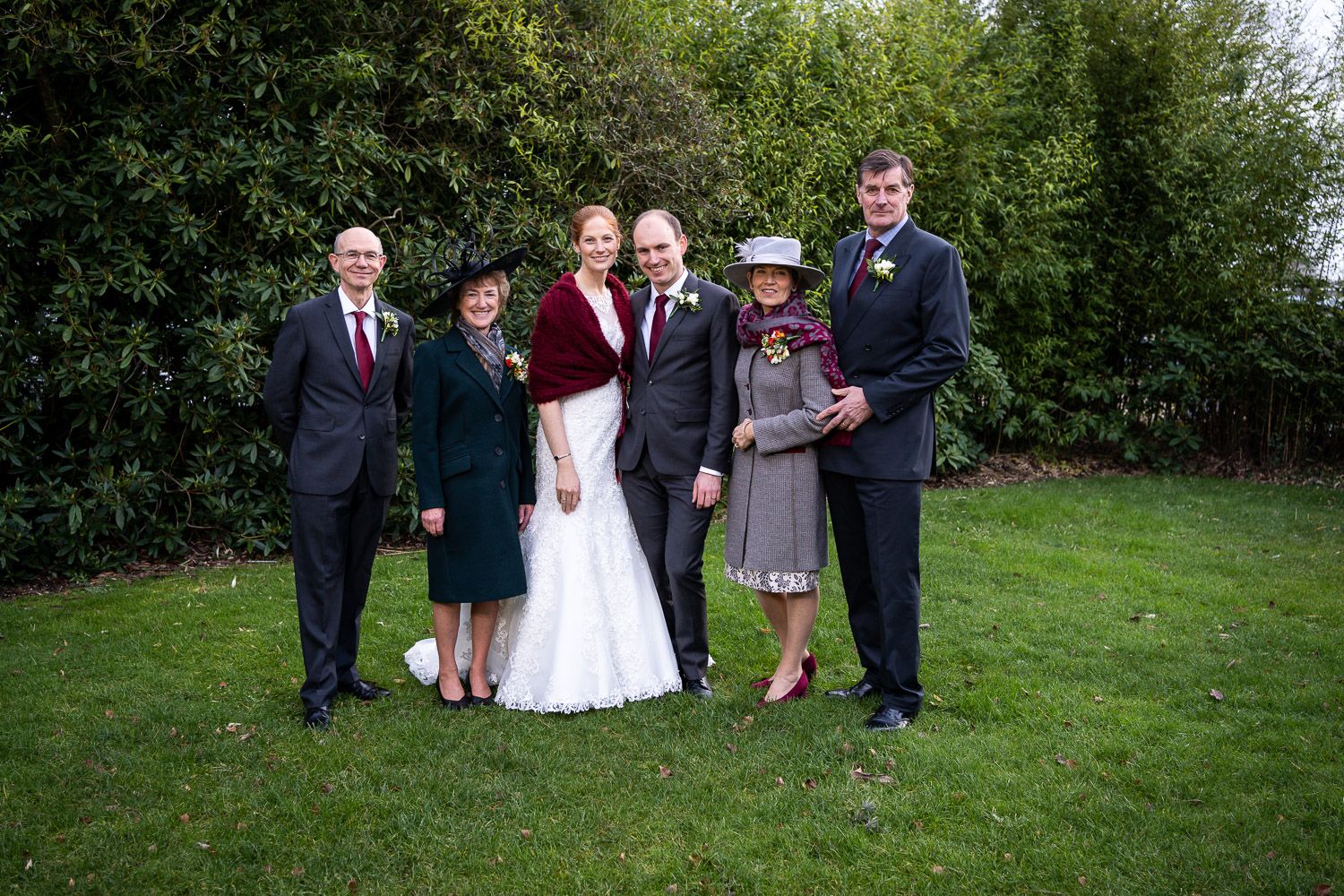 A formal group poses together outdoors on a green lawn with hedges in the background during a wedding celebration.