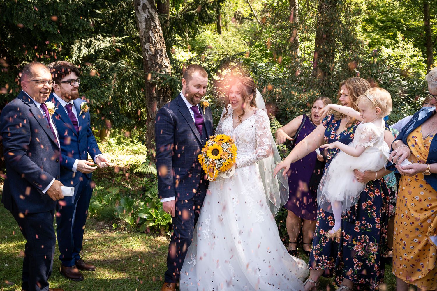 Wedding guests shower the happy couple with confetti during a sunny outdoor celebration with sunflowers and floral decor.