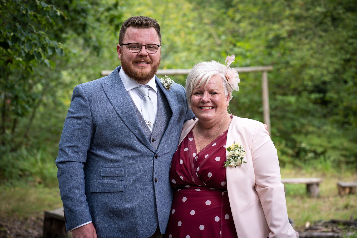 A couple stands together outdoors in formal attire with a gray suit and pink cardigan against a green forest backdrop.