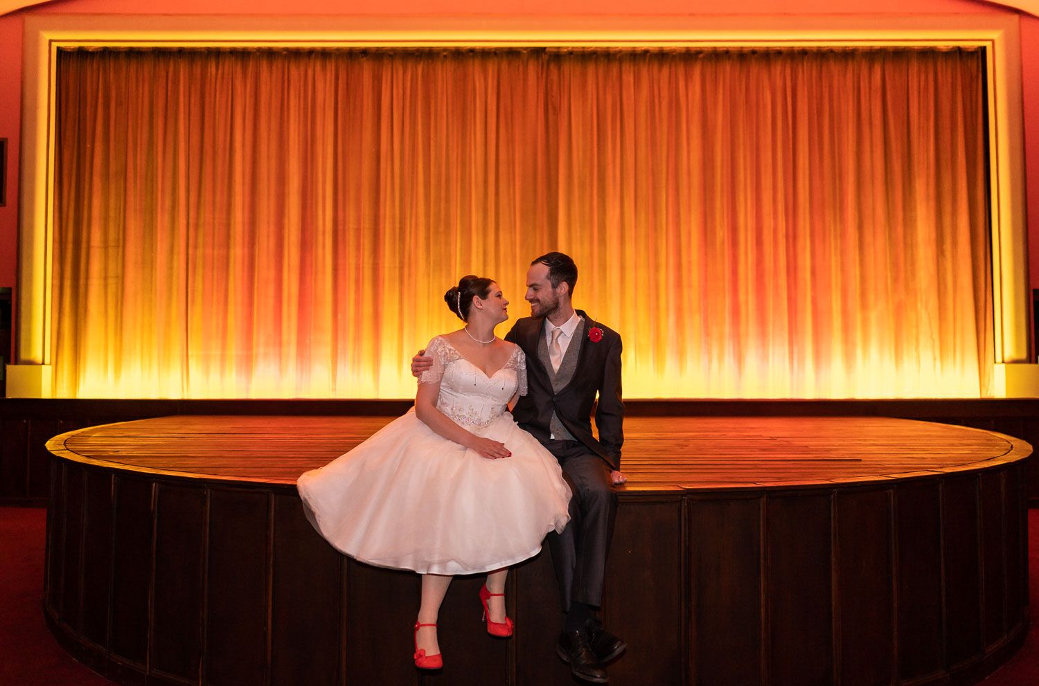 A couple in formal attire shares a romantic moment on an illuminated theater stage with orange curtains.