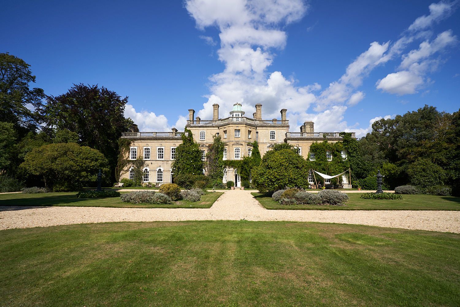 Elegant English manor house with manicured lawn and gardens on a sunny day with blue skies and white clouds.