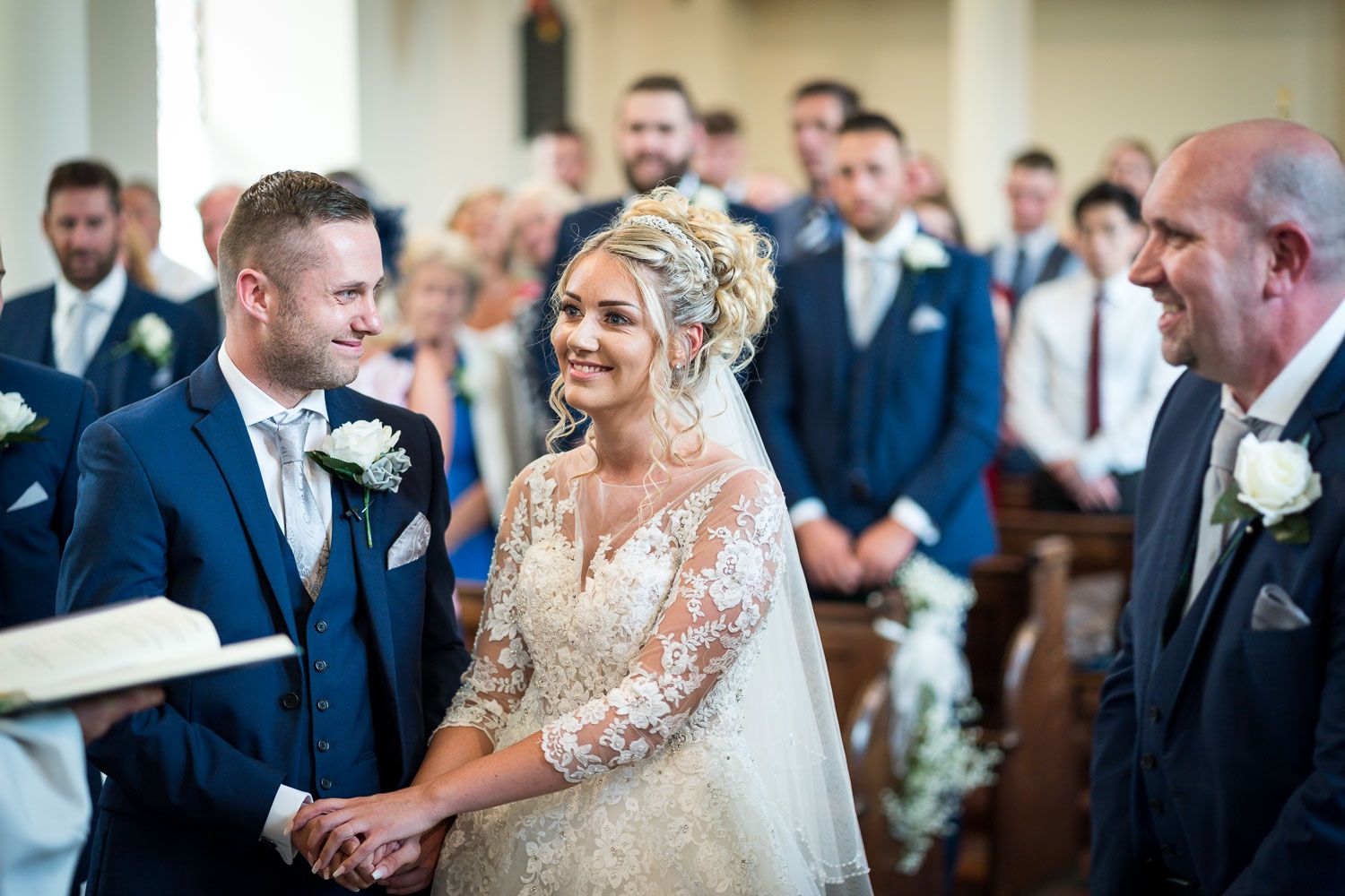 A joyful wedding ceremony moment in a church with the couple exchanging rings while guests in navy suits look on.