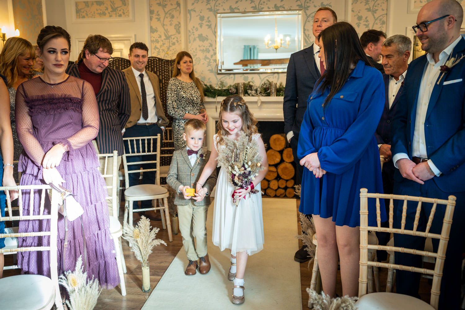 Wedding ceremony guests watch as two small children walk down the aisle in an elegant indoor venue.