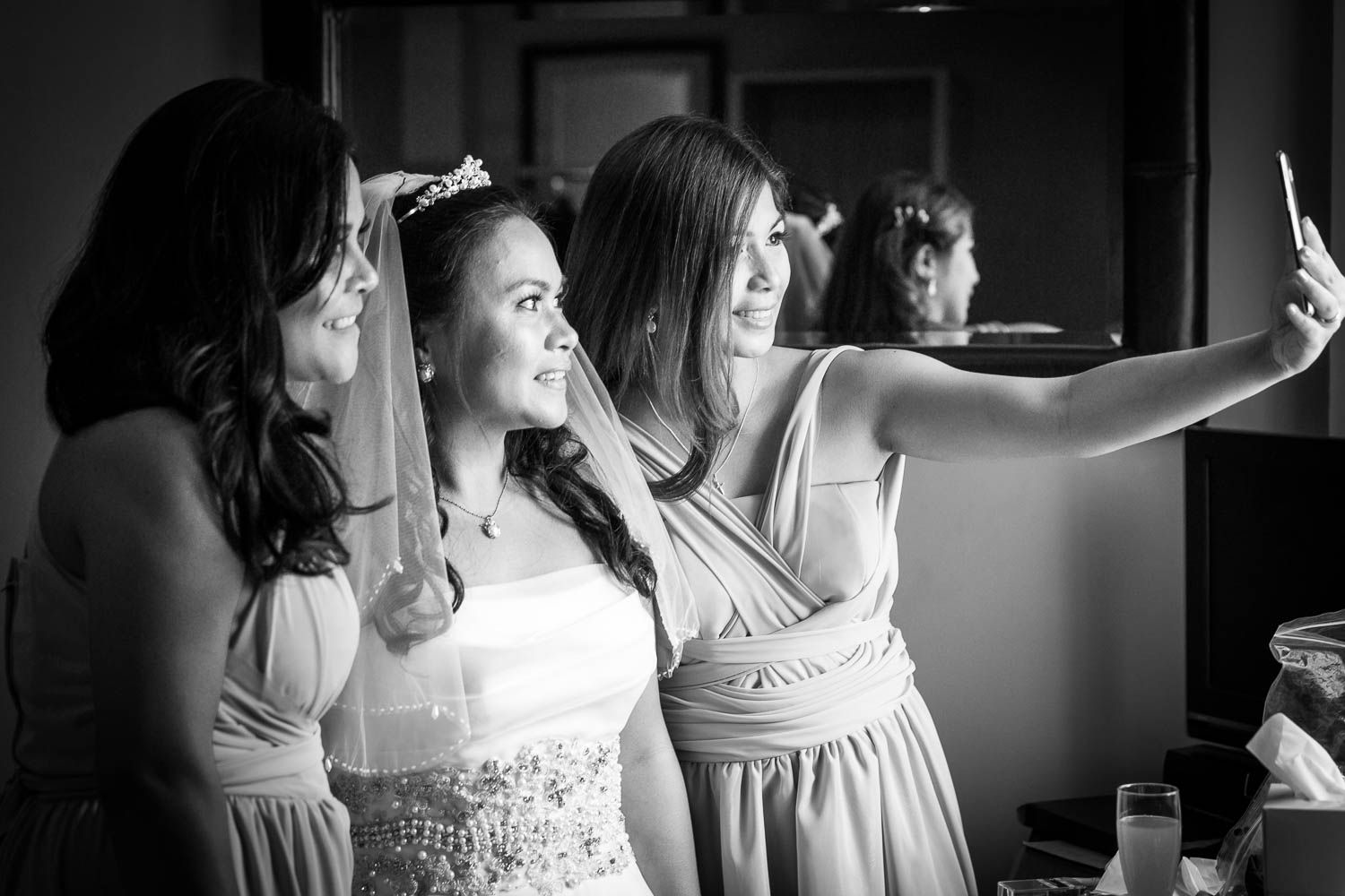 A bride and bridesmaids in light-colored dresses take a selfie together in black and white photography.