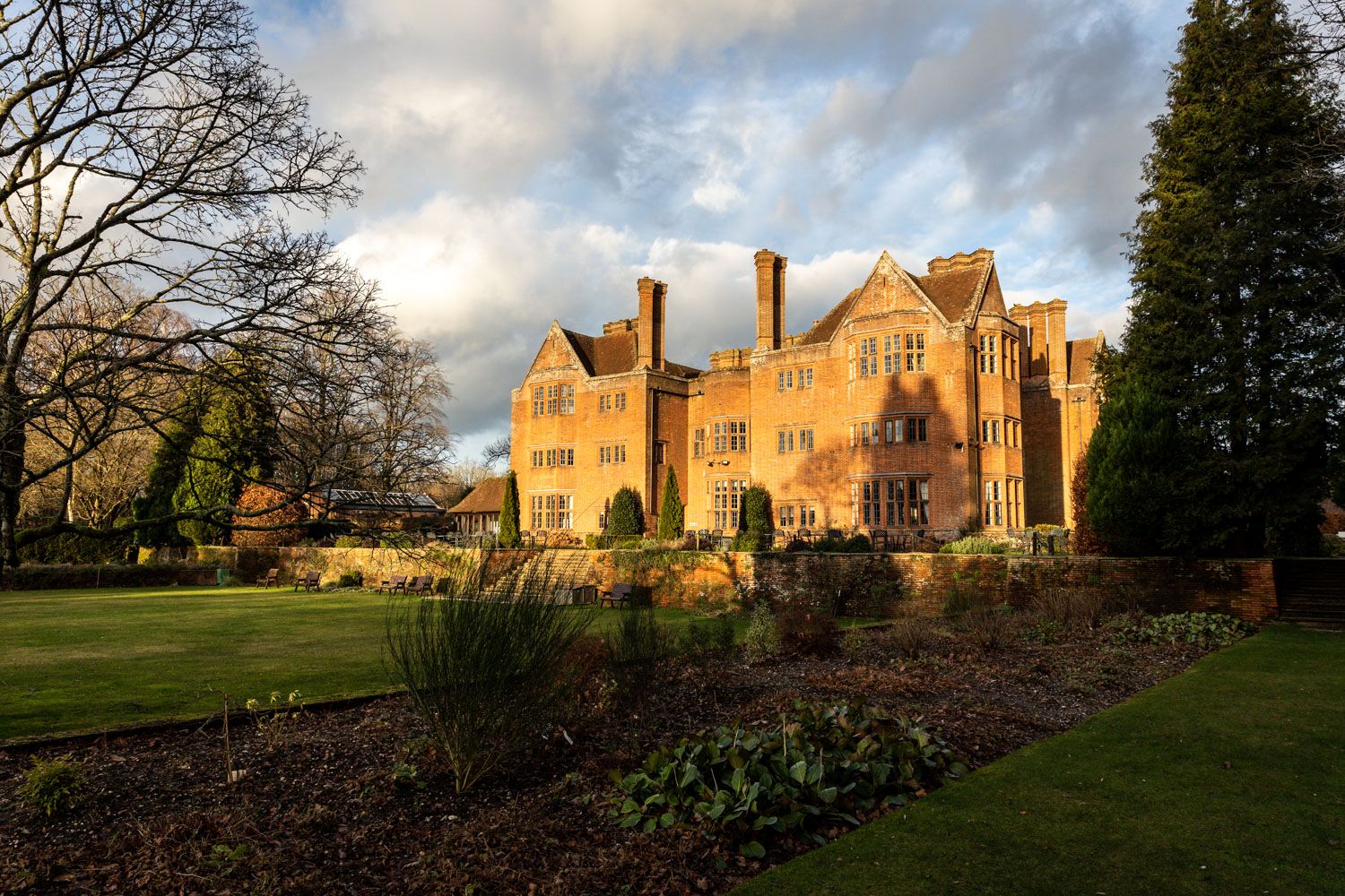 Historic English manor house with red brick facade illuminated by golden sunset light across manicured garden grounds.