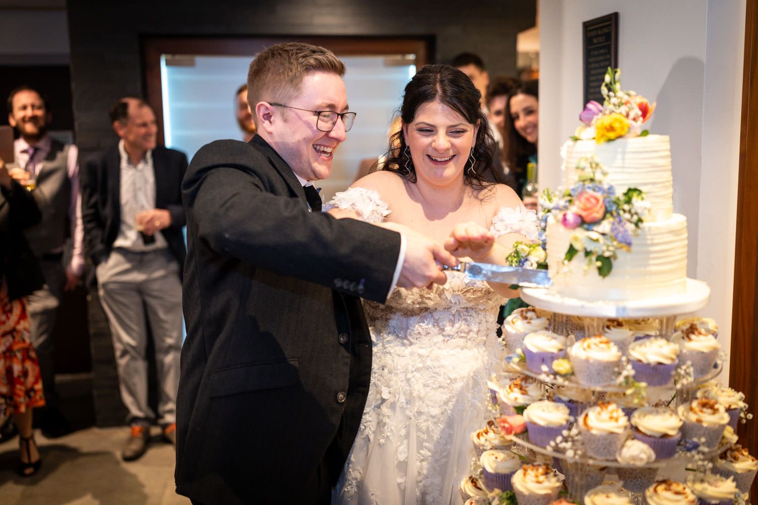 A joyful couple cuts their wedding cake together at their reception while guests look on and smile.