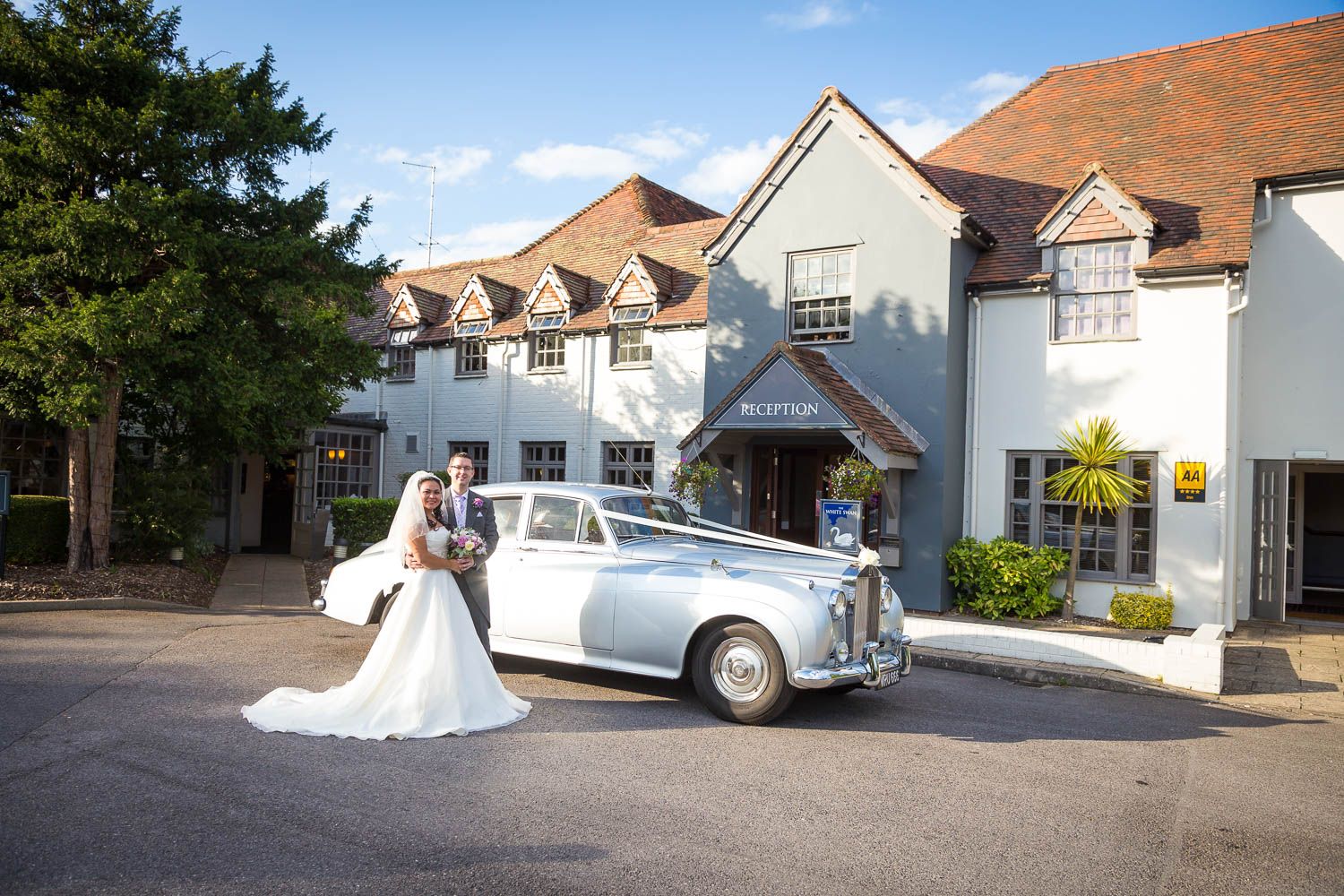 A classic white vintage Bentley car parked in front of a white Tudor-style building on a sunny day.