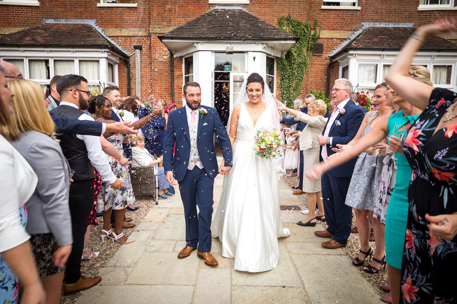 Wedding guests line a walkway throwing confetti as the newlywed couple exits against a backdrop of red brick buildings.