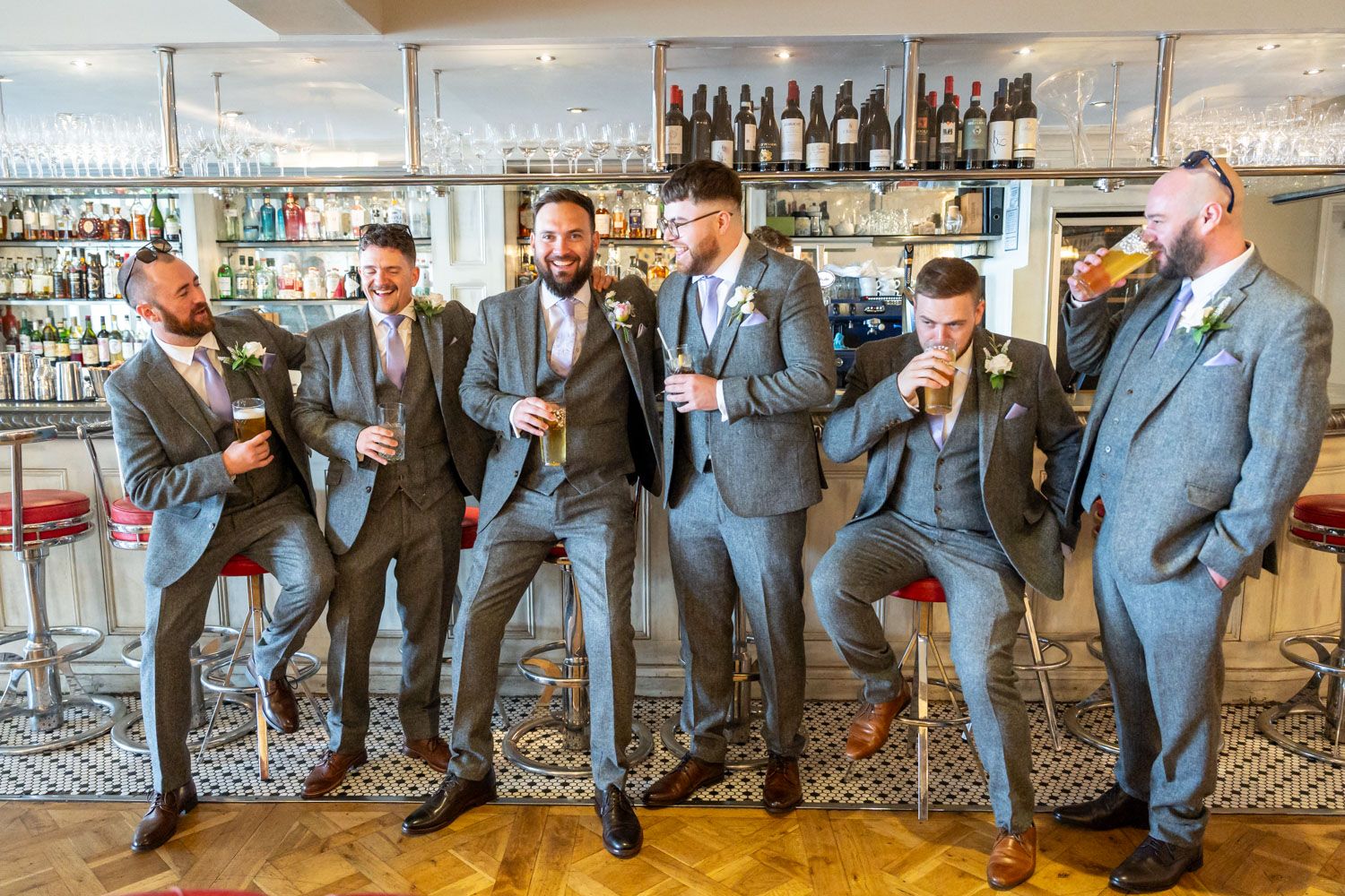 Six groomsmen in grey suits laughing and celebrating with drinks at a bar before a wedding ceremony.