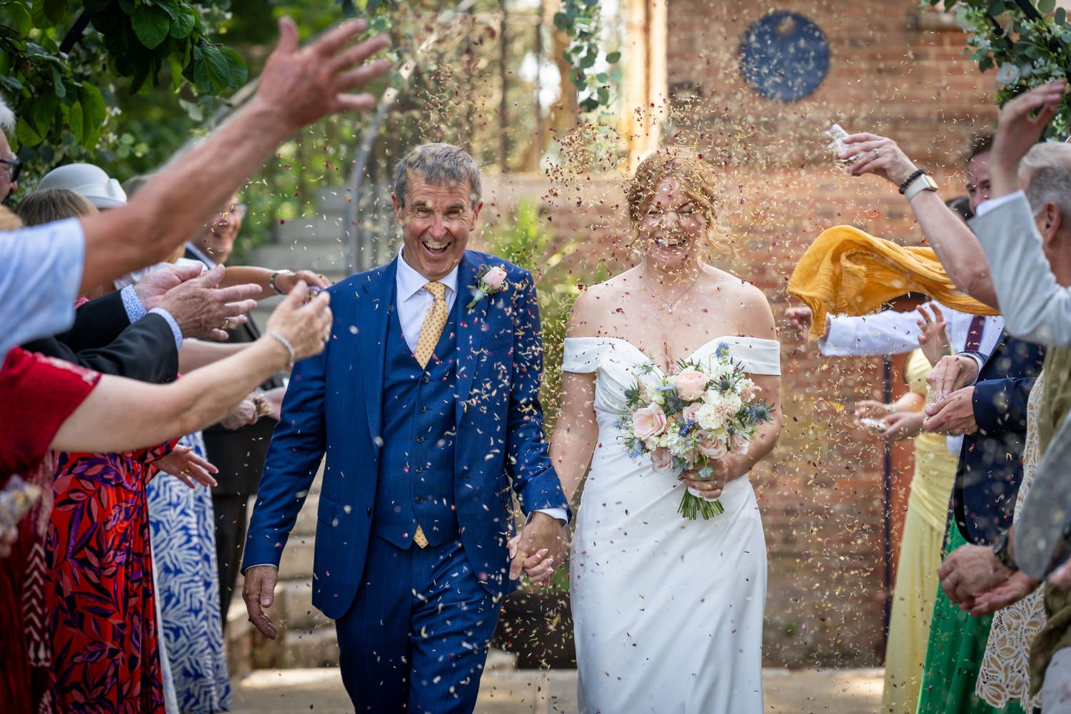 Joyful newlyweds walking through a confetti shower thrown by cheering wedding guests outdoors.
