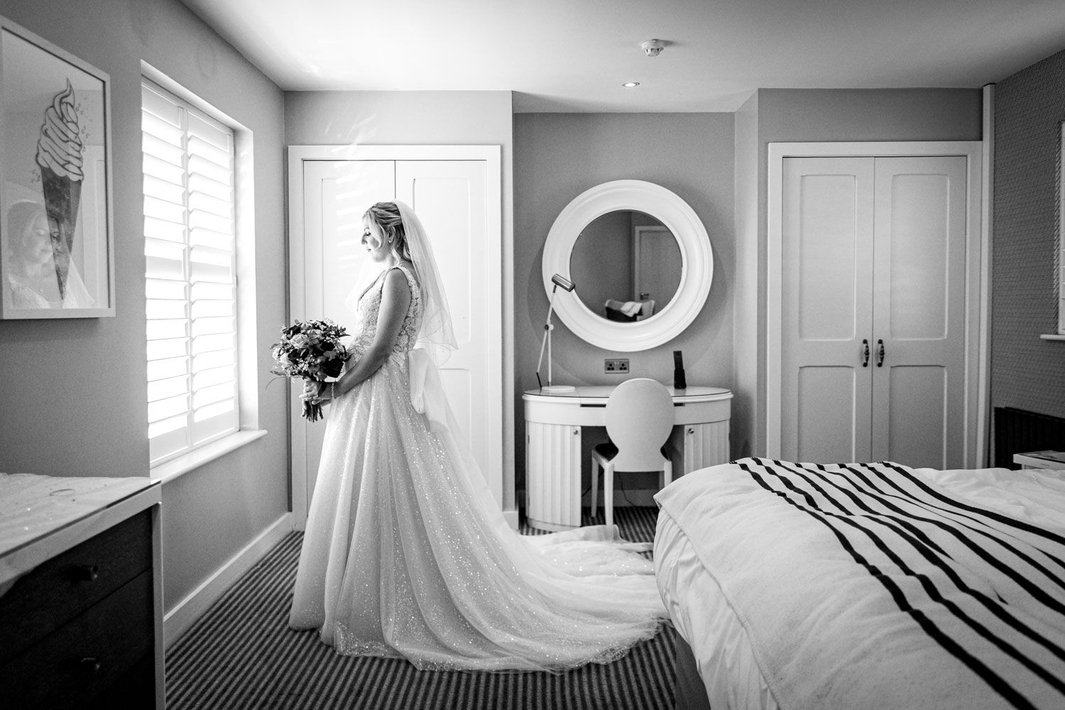Elegant bride in a long white gown standing by a window in a black and white photo.