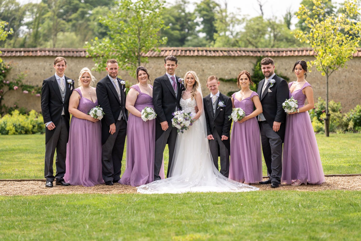 Wedding party in formal attire poses outdoors with bridesmaids wearing flowing lavender dresses against a garden wall.