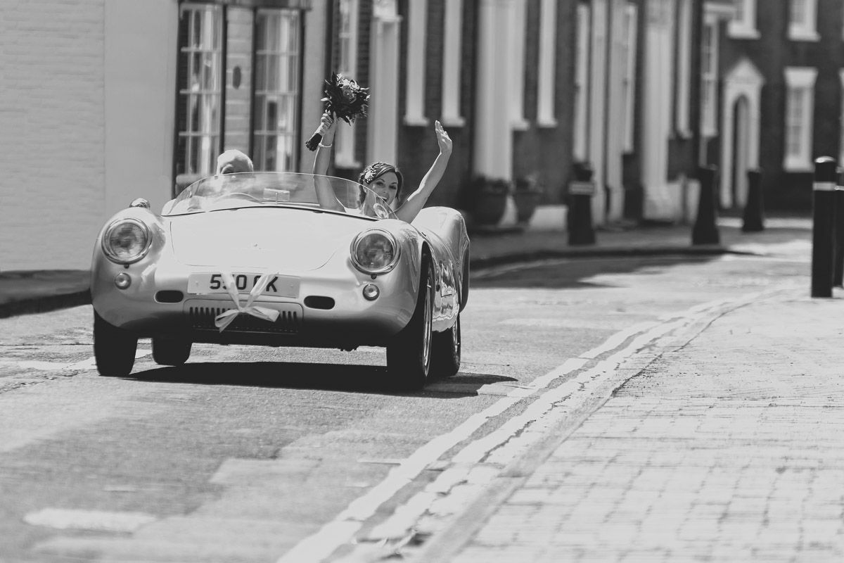 A classic Porsche 550 Spyder drives down a narrow cobblestone street in black and white photography.