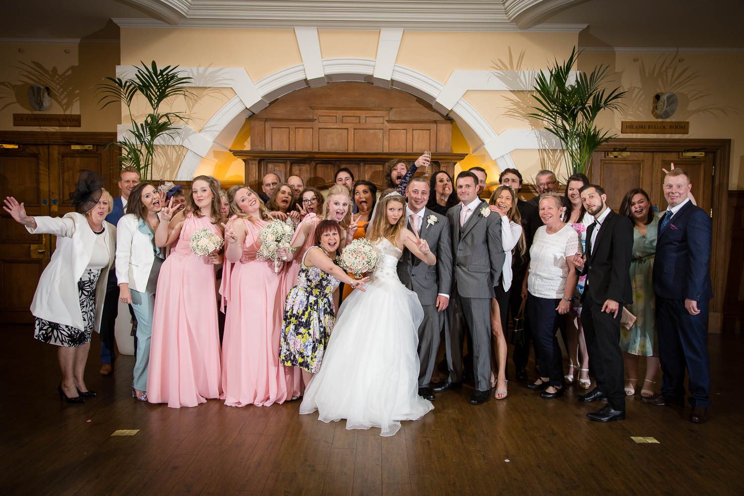 Wedding party and guests pose together in a grand ballroom with an elegant arched entrance and palm plants.