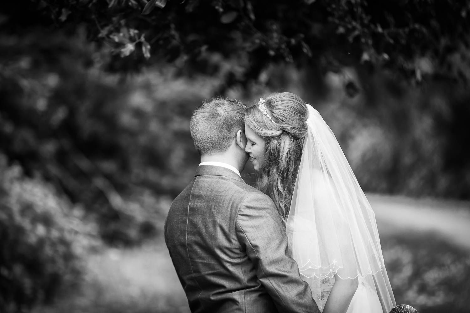 A black and white wedding photo shows a bride in a veil resting her head on the groom's shoulder outdoors.