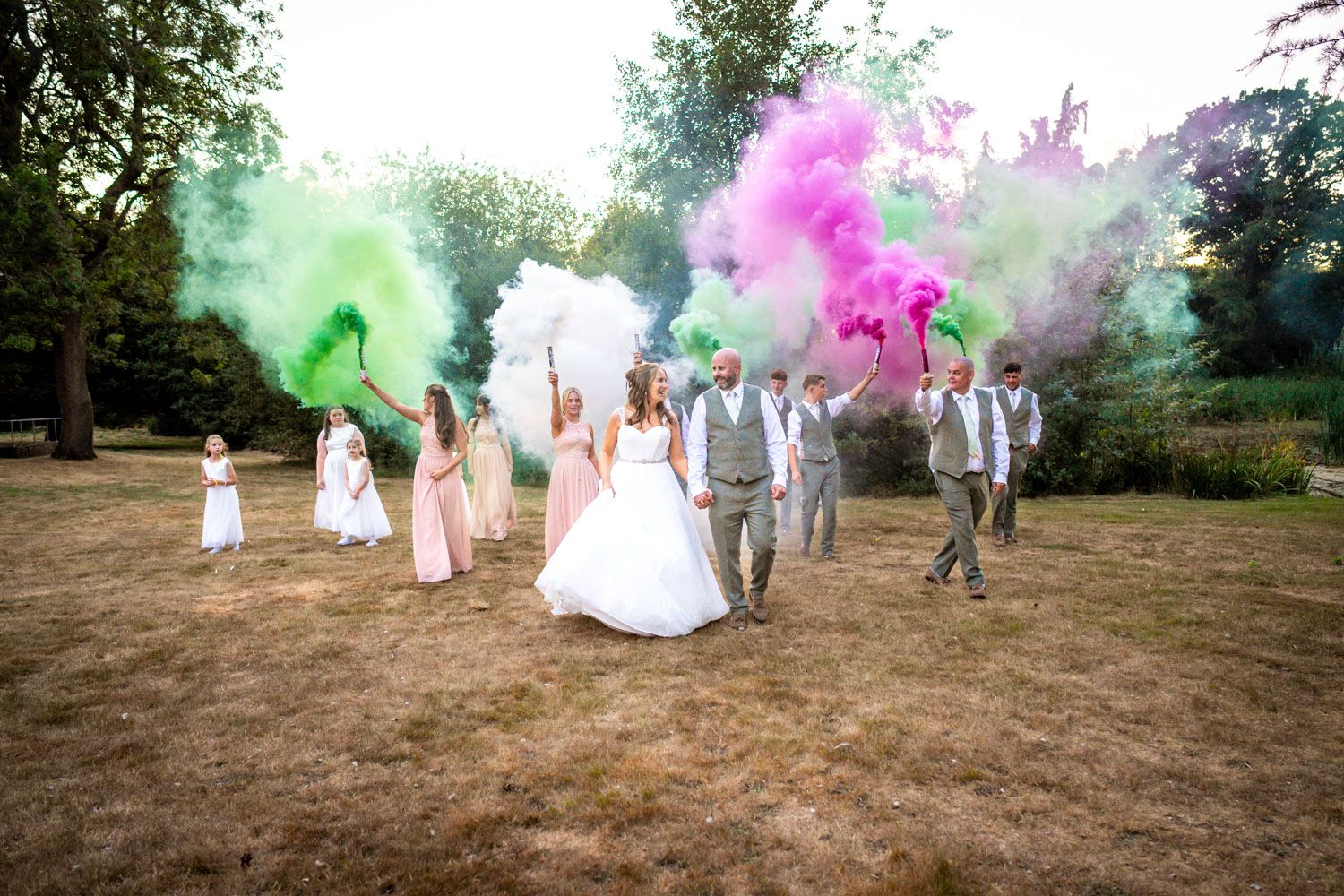 Wedding party celebrating outdoors with colorful pink, green, and purple smoke bombs on a sunny day.