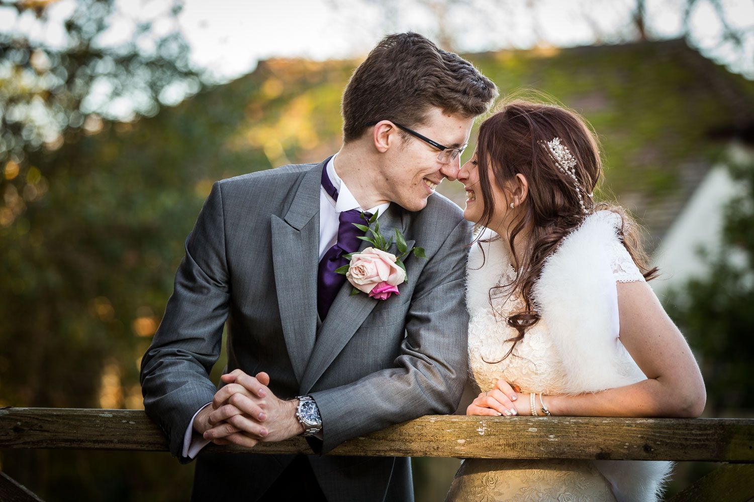 A romantic wedding moment shared between newlyweds in formal attire leaning on a wooden railing at sunset.