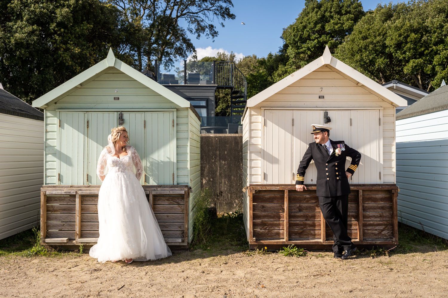 Two colorful pastel beach huts with couple in wedding attire standing outside them on sandy beach.