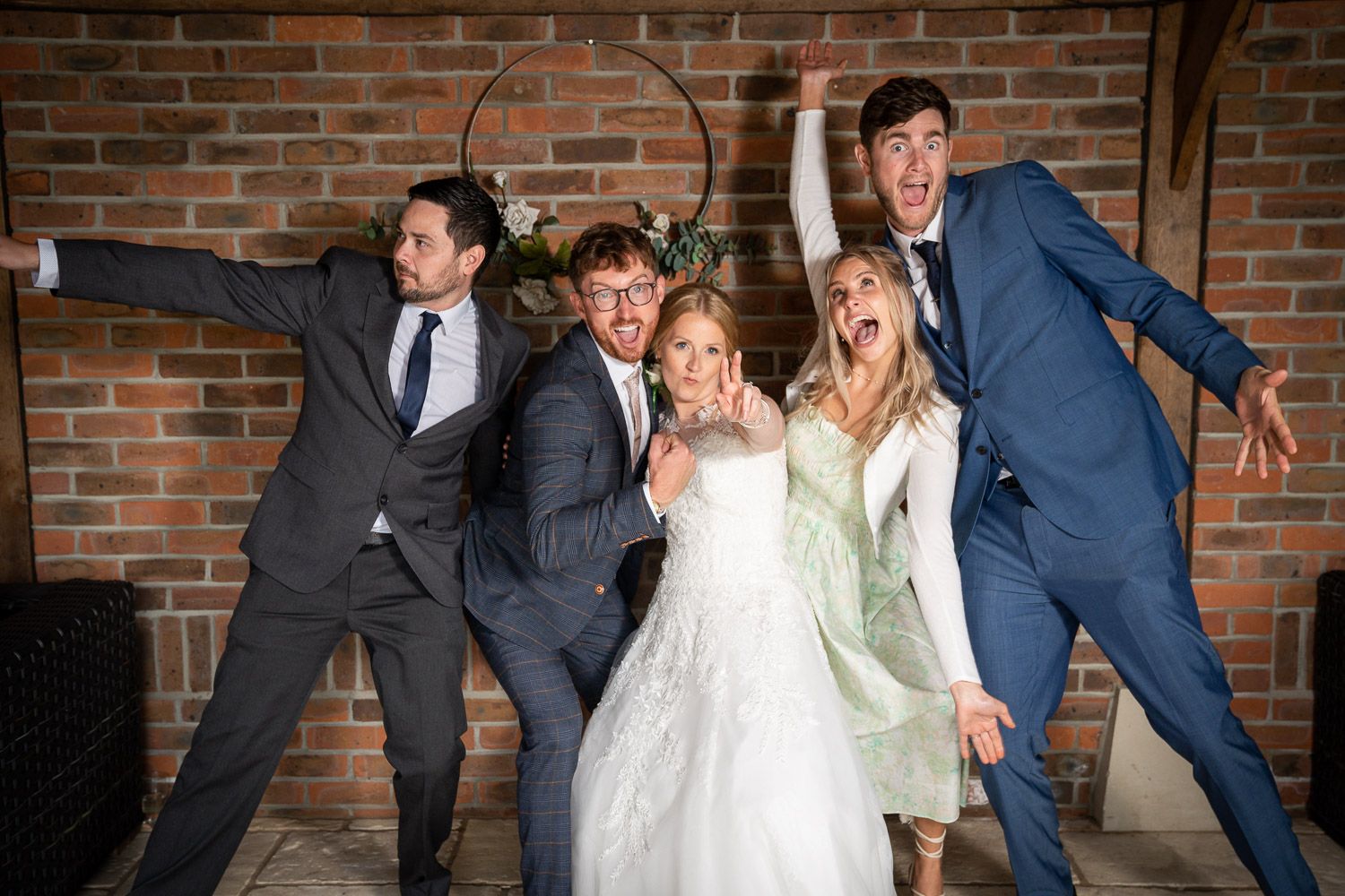 Wedding party poses playfully against brick wall in mix of formal suits, white dress and sage colored dress.