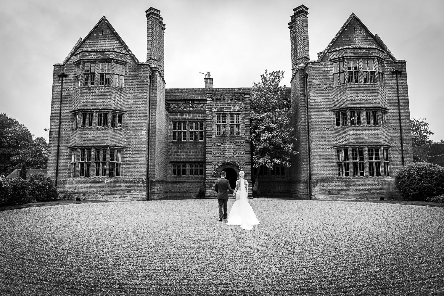 Black and white wedding photo of a couple standing in front of a grand stone manor with twin towers.