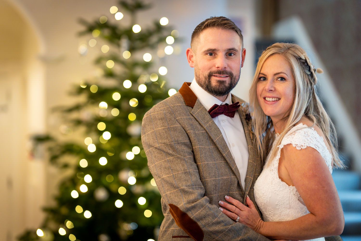 A bride and groom pose together in front of a glowing Christmas tree during their winter holiday wedding celebration.