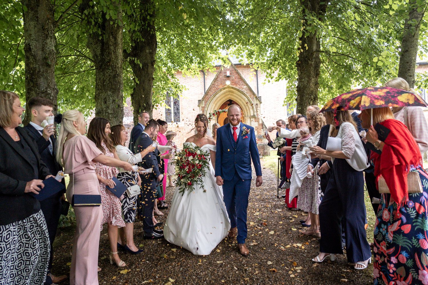 Wedding guests lining a tree-covered path throw confetti as newlyweds walk through after church ceremony.