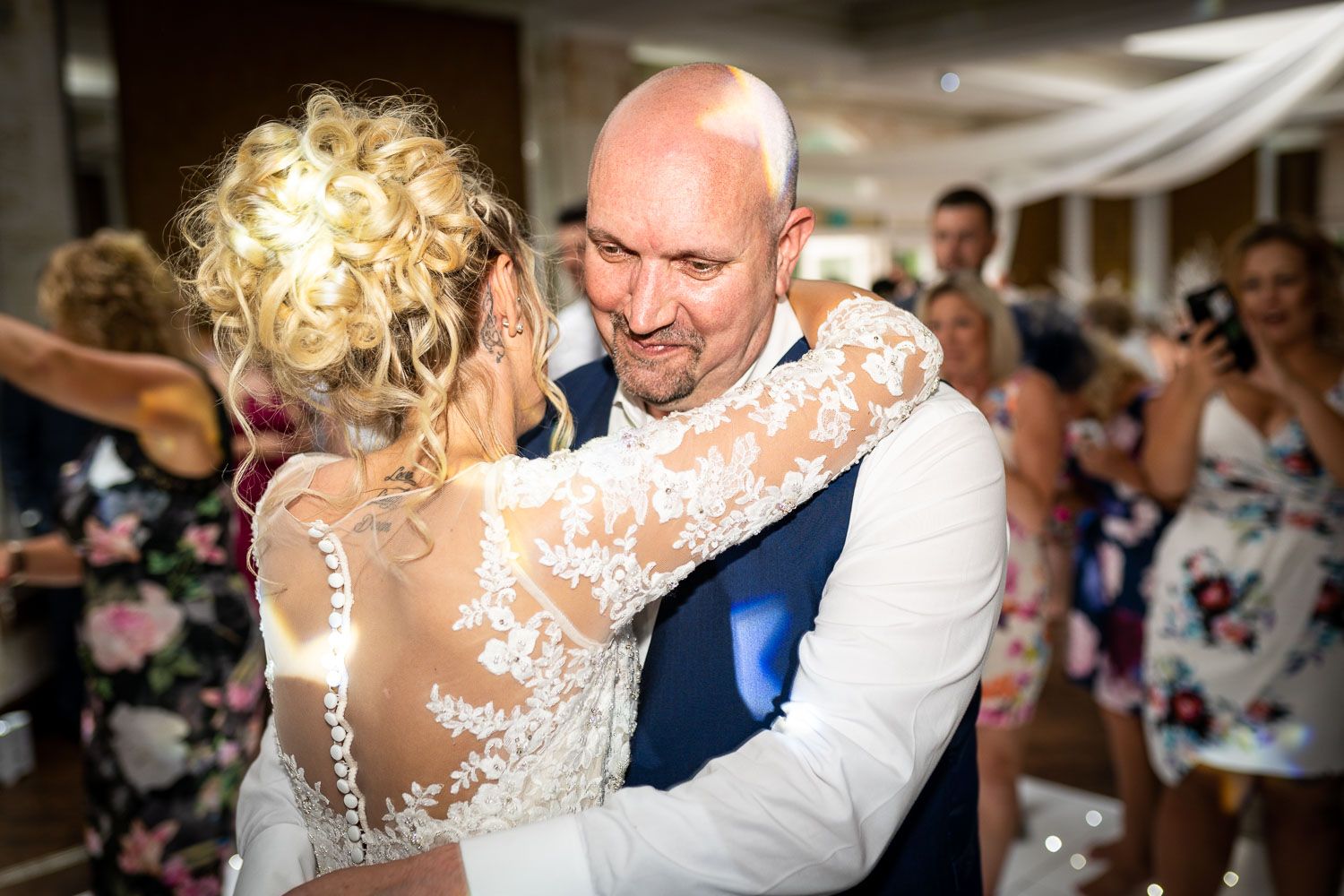 Couple shares a tender embrace during a wedding celebration while guests look on in the background.