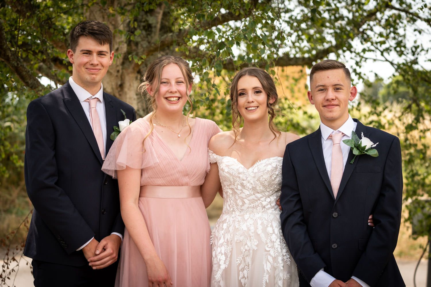 Two couples dressed formally in wedding attire including navy suits, pink ties, and a lace white wedding gown pose outdoors.