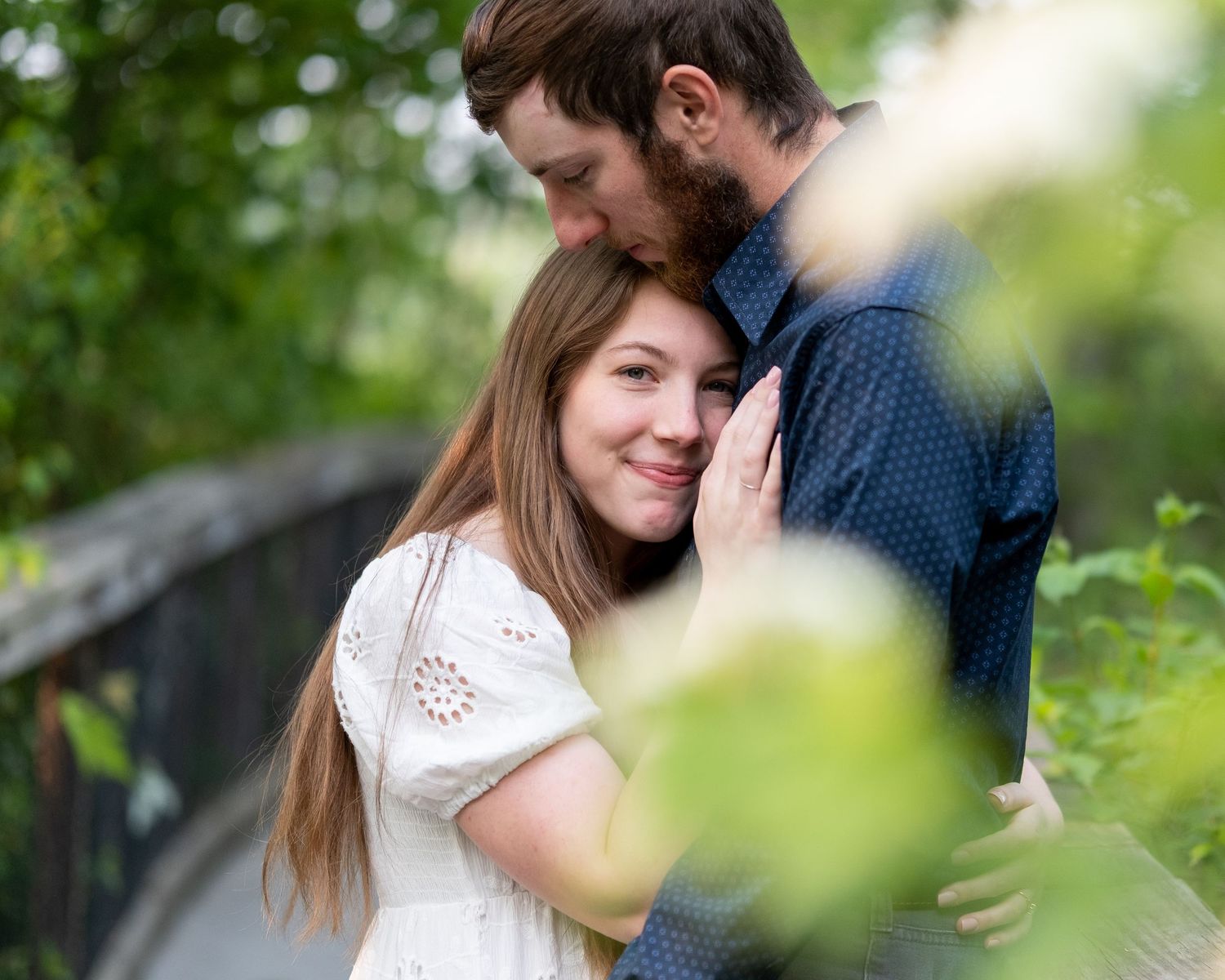 A smiling woman with long hair leans into a bearded man in a blue shirt as they embrace outdoors among green foliage.